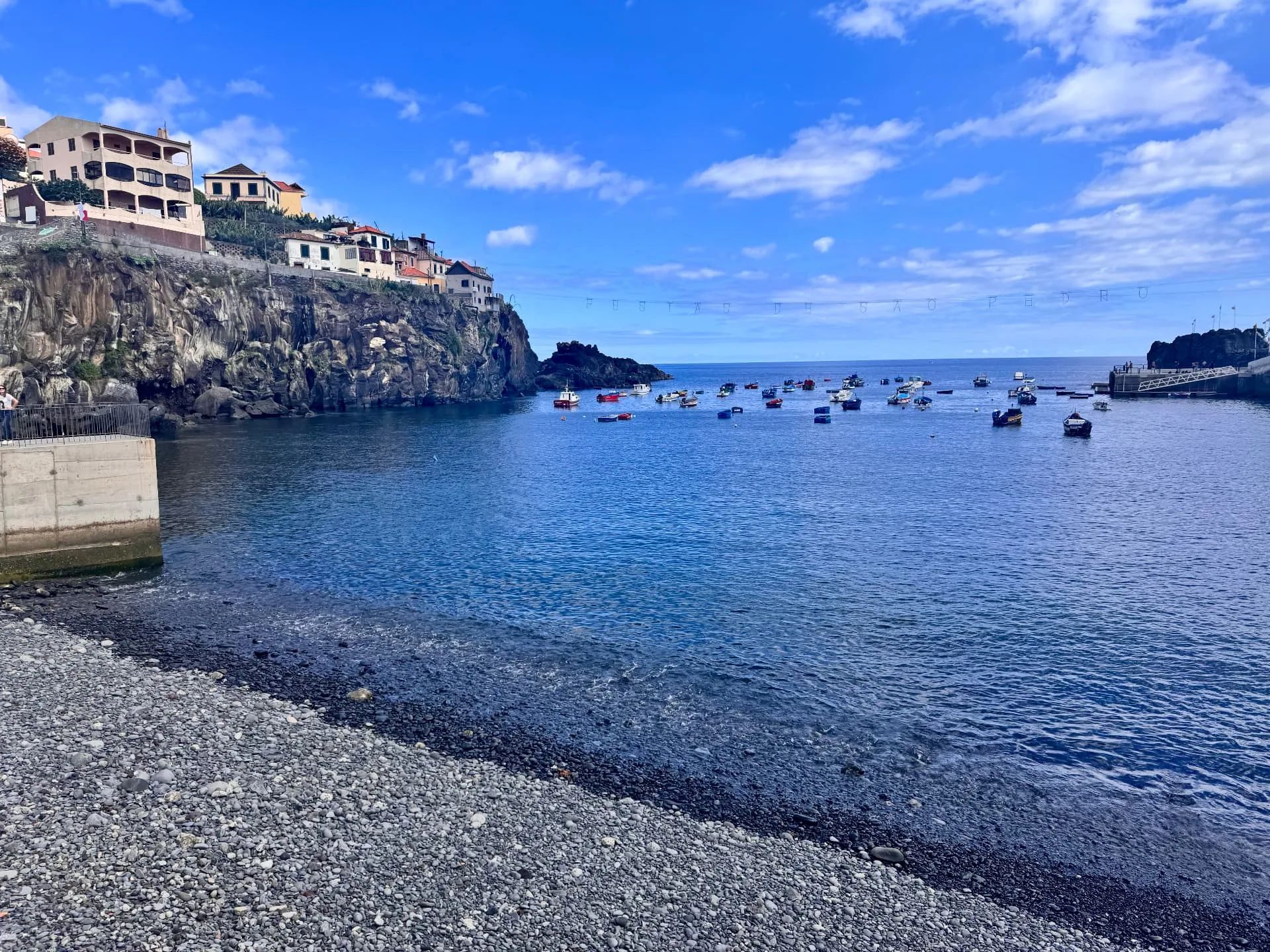 Praia de calhau e baía azul e calma em Câmara de Lobos, com casas ao longo da falésia de basalto e barcos de pesca coloridos
