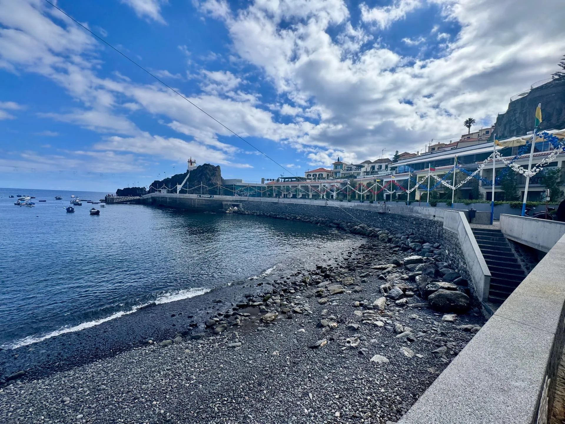 Praia de calhau e muralha em Câmara de Lobos, com grinaldas de festa ao longo dos edifícios da marginal e o farolim no rochedo