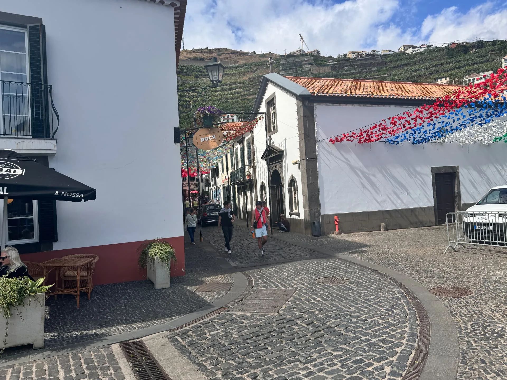 Rua empedrada junto a uma capela branca em Câmara de Lobos, com grinaldas de flores de papel e socalcos de bananeiras na encosta