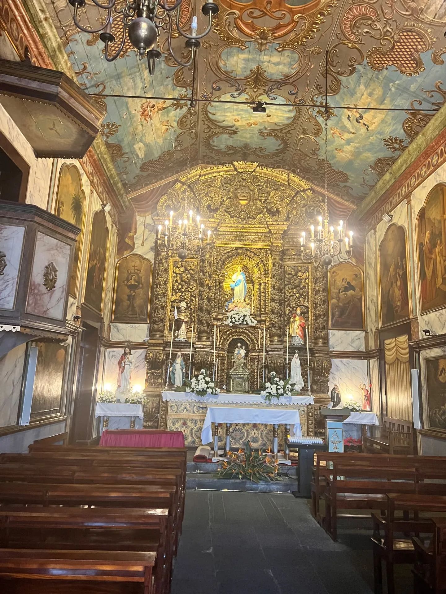 Interior de uma igreja em Câmara de Lobos: teto de madeira pintado, pinturas nas paredes, bancos de madeira e retábulo dourado