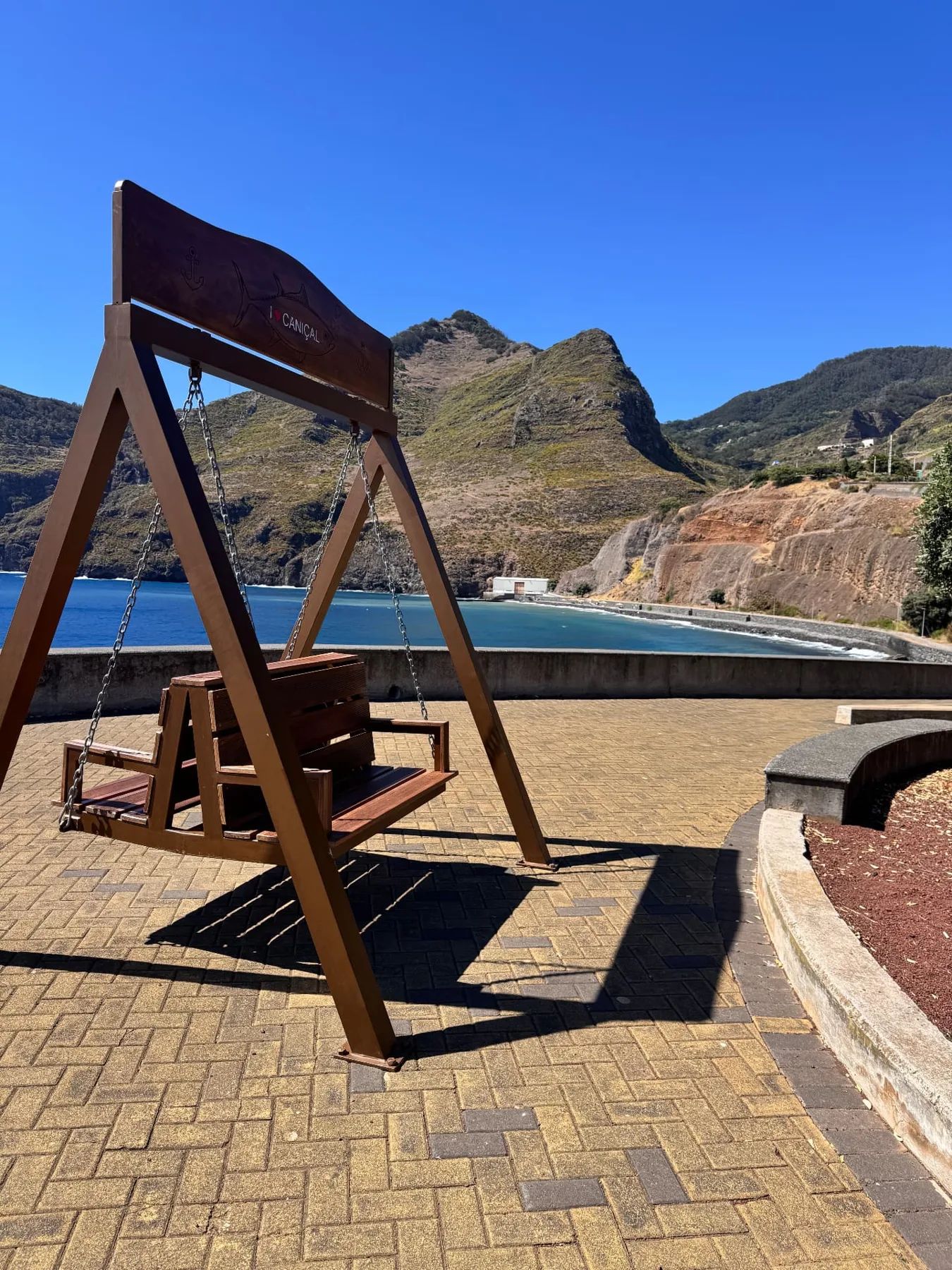 Balanço de madeira com a inscrição «I love Caniçal» no passeio marítimo, com a baía e as montanhas ao fundo