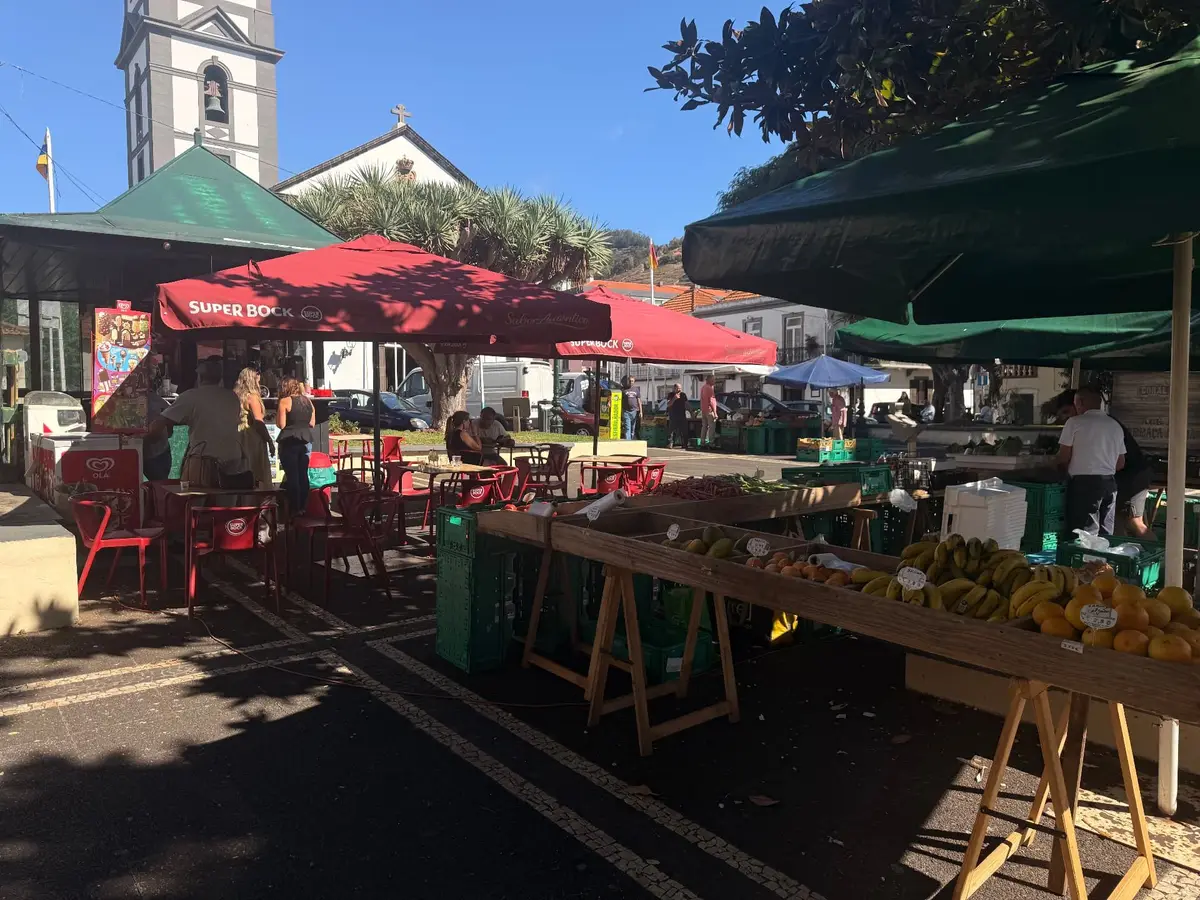 Farmers market stalls under red and green parasols in Caniço town square with the church bell tower behind, Madeira