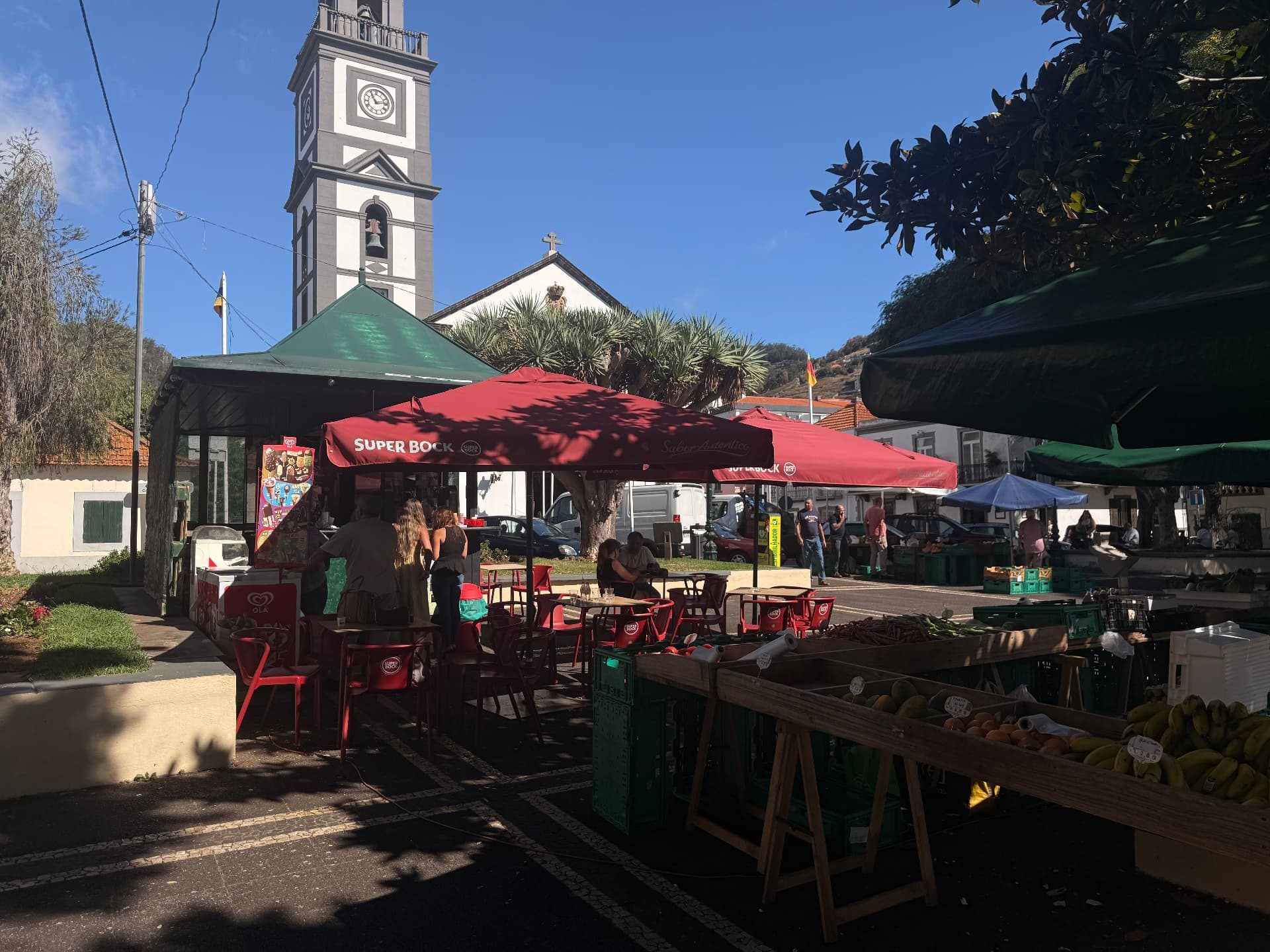 Market stalls and a cafe kiosk in Caniço square beneath the white clock tower of the parish church