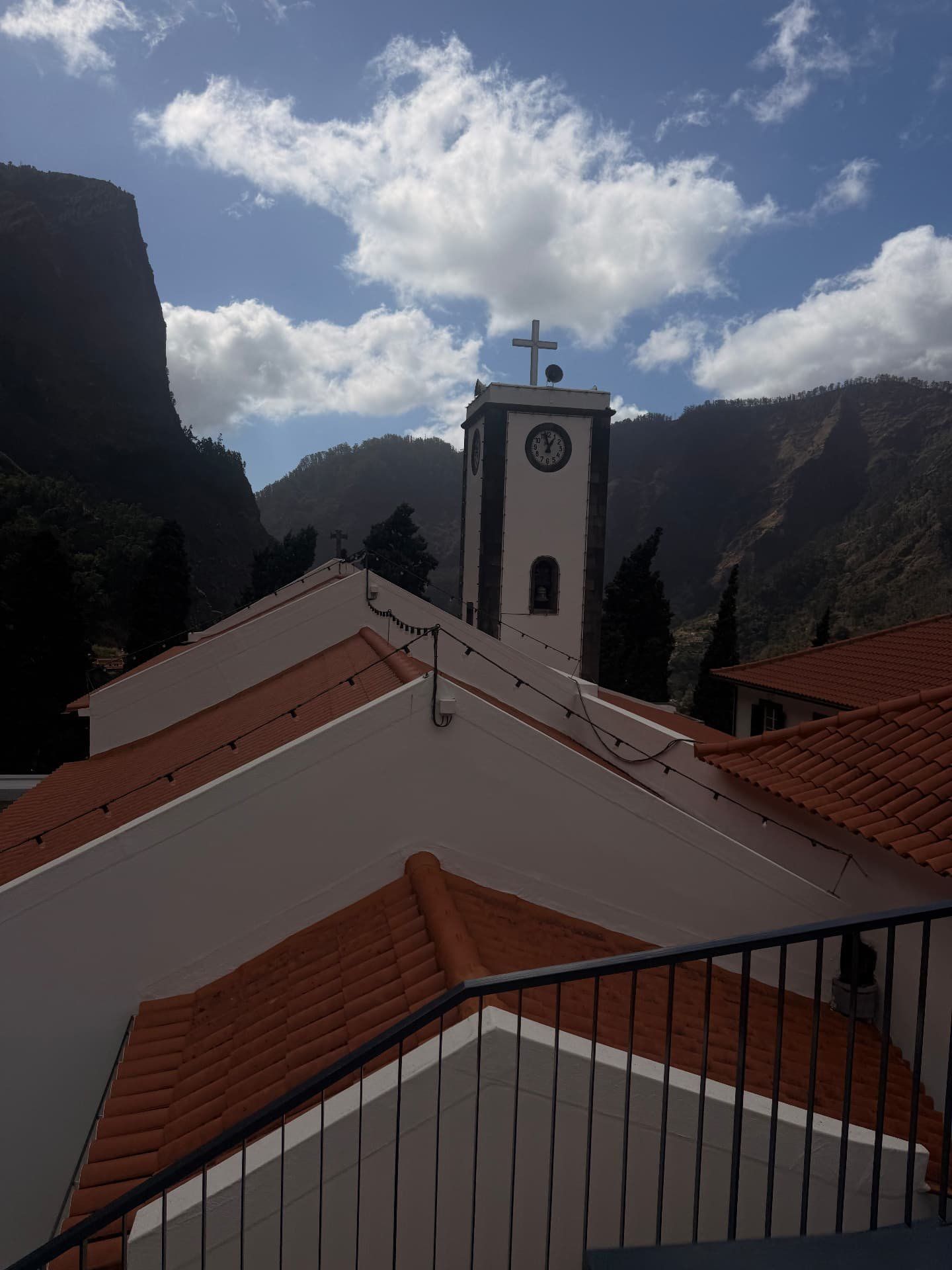 Torre branca do relógio da igreja paroquial do Curral das Freiras sobre telhados vermelhos, com as encostas do vale atrás