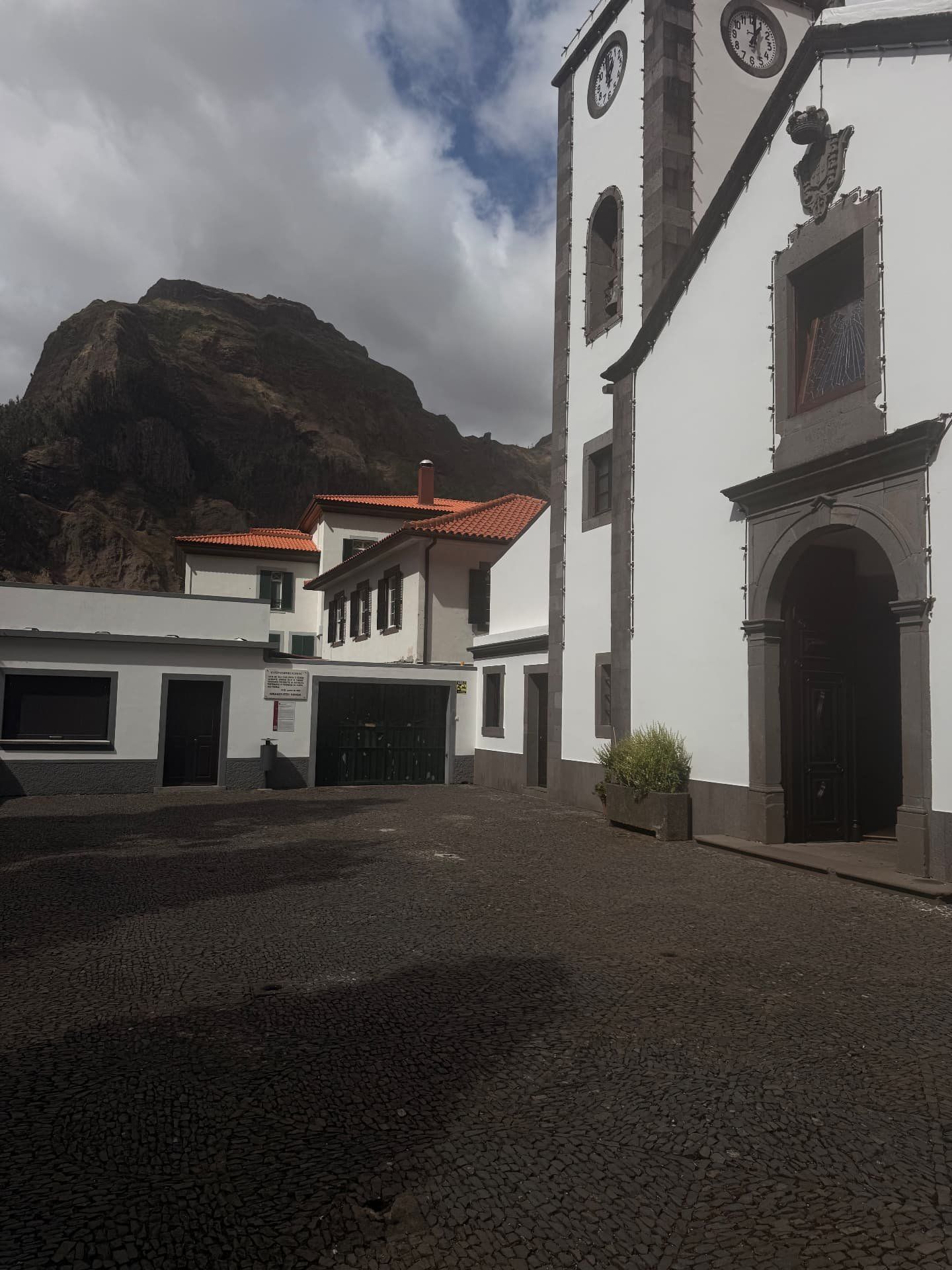 Adro calcetado e casa paroquial junto à igreja, com um pico rochoso ao fundo