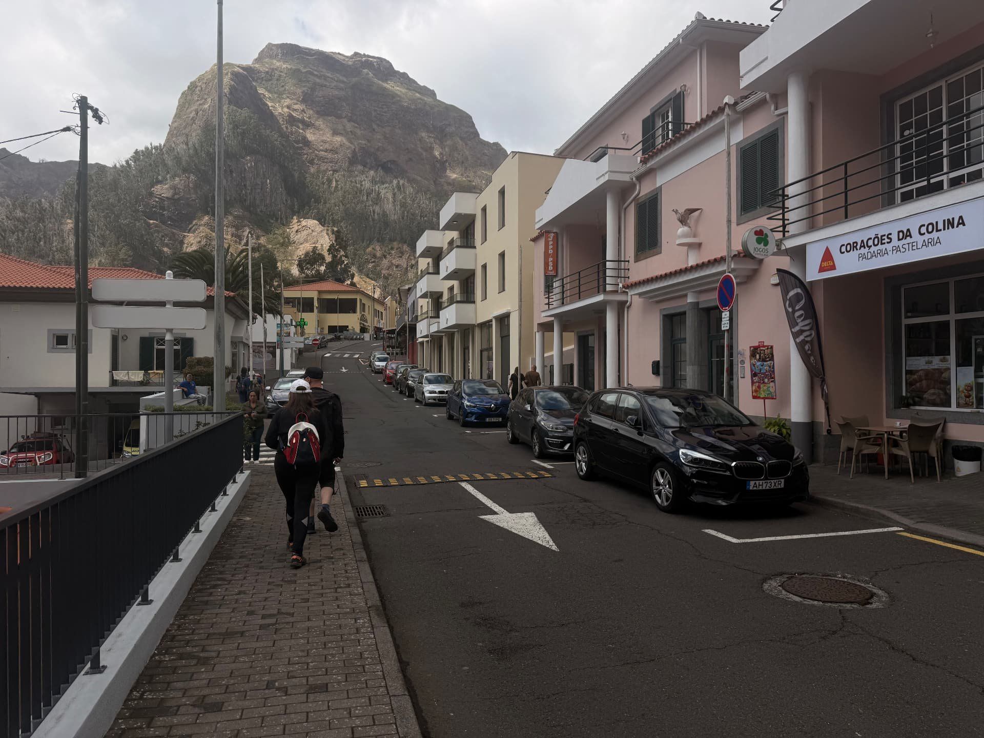 Rua principal do Curral das Freiras, com uma padaria e carros estacionados, por baixo de um enorme pico rochoso