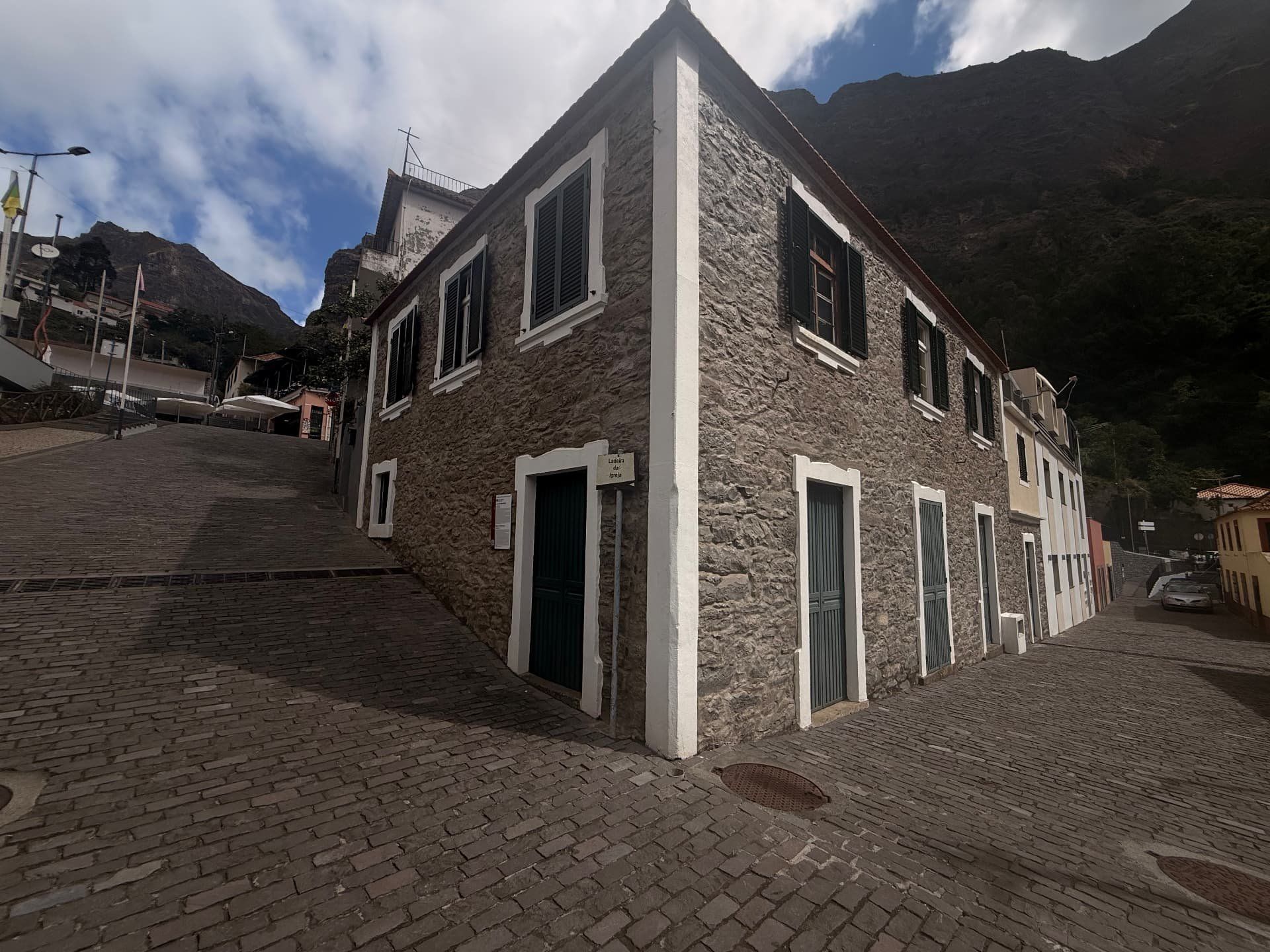 Casa de pedra de dois pisos com cantarias brancas e portadas verdes numa esquina de calçada no Curral das Freiras