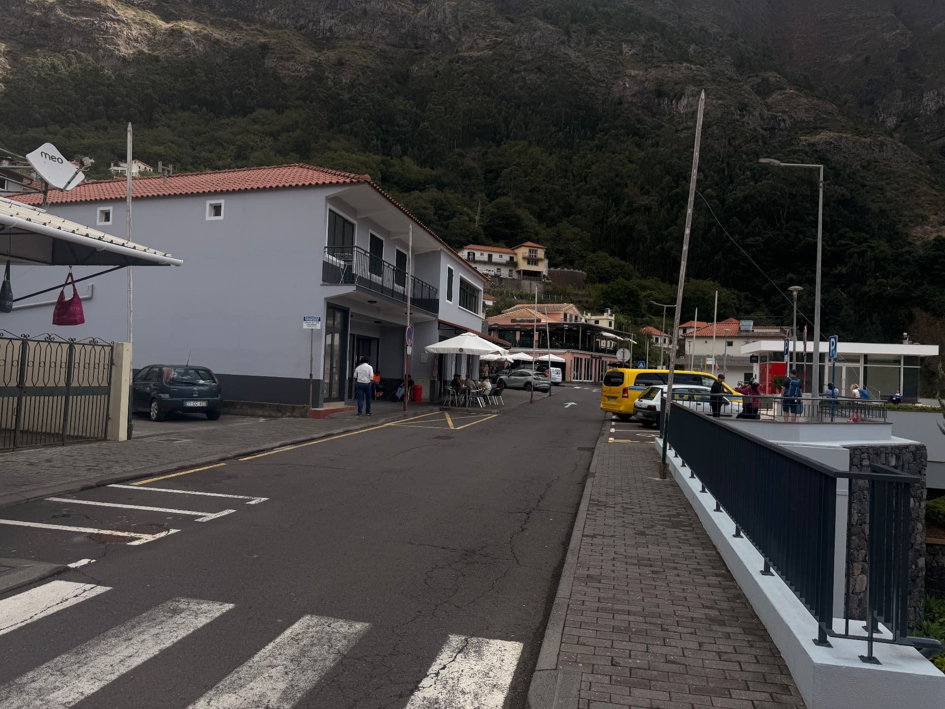 Centro da aldeia do Curral das Freiras com esplanadas, um parque de estacionamento e a encosta arborizada atrás