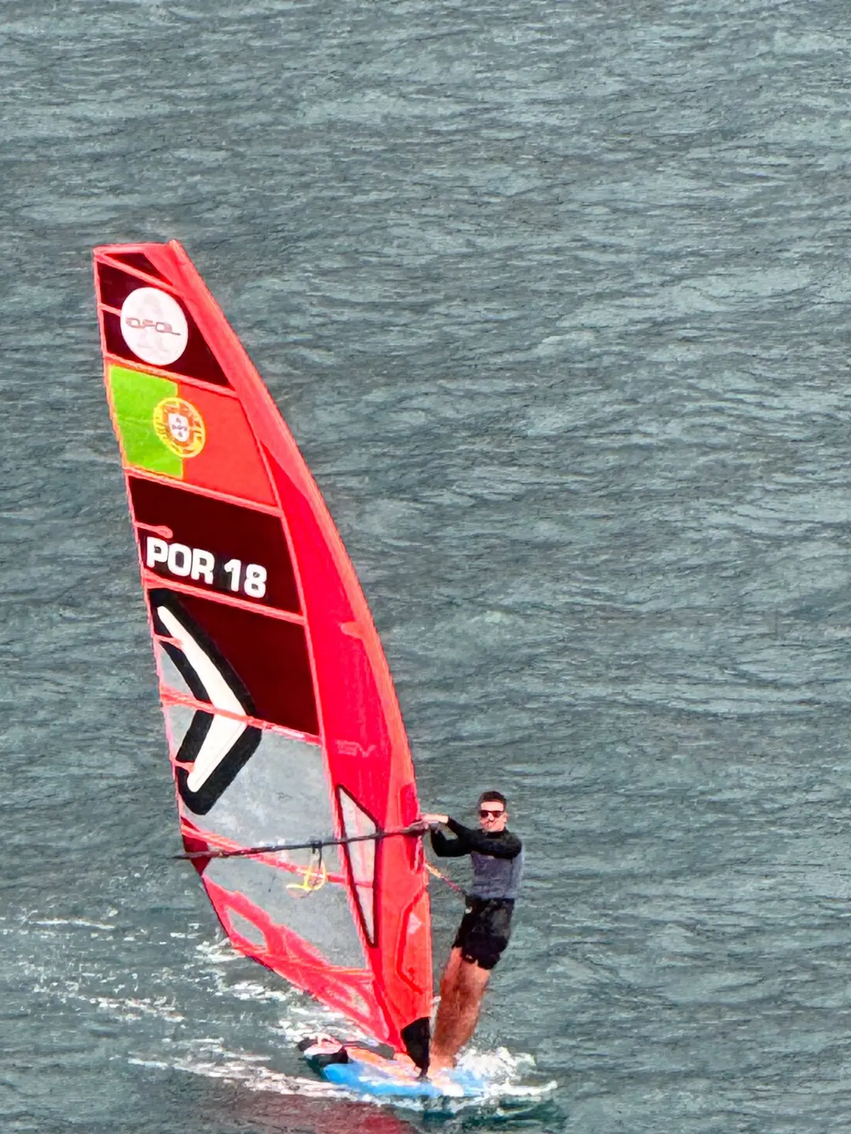 A windsurfer with a red Portuguese-marked sail crossing Funchal Bay with the city and mountains behind, Madeira