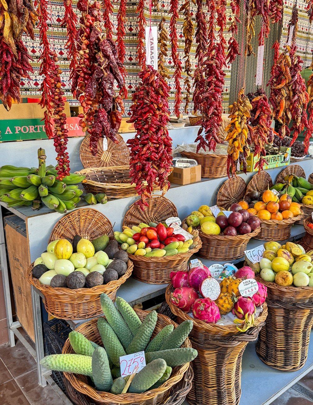 Strings of dried red chillies hanging above wicker baskets of pitaya, custard apples, tomarillos and prickly pears at Mercado dos Lavradores, Funchal, Madeira