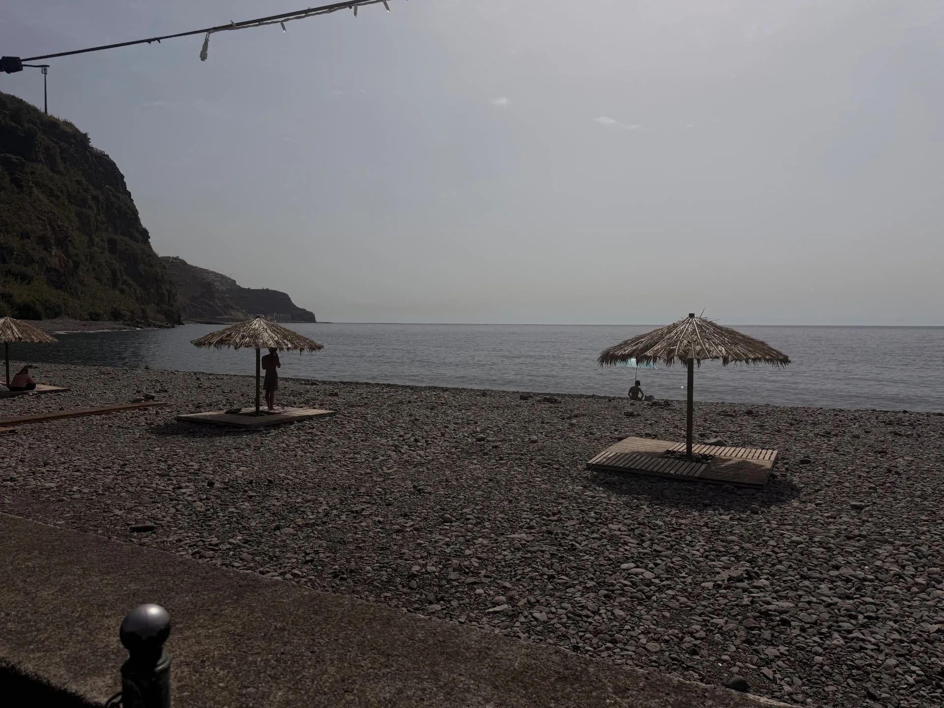 Pebble beach at Lugar de Baixo with thatched parasols on wooden platforms and the coast running west