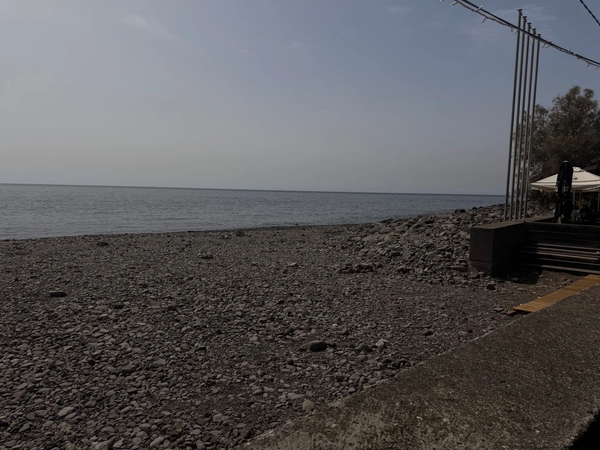 Wide pebble shore and calm sea at Lugar de Baixo, with a beach bar terrace on the right