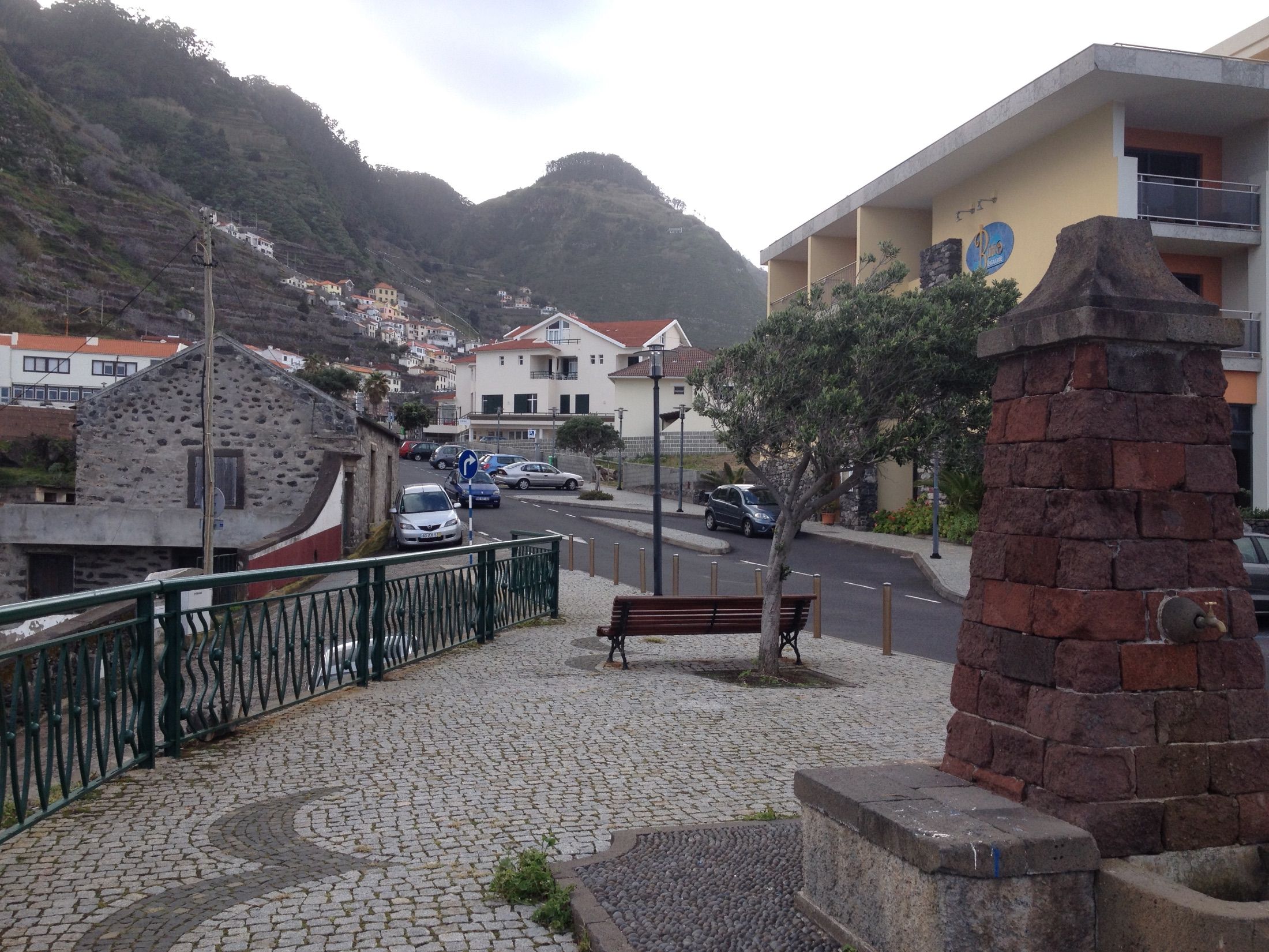 Chafariz de pedra e banco junto a uma rua sob encostas íngremes com socalcos