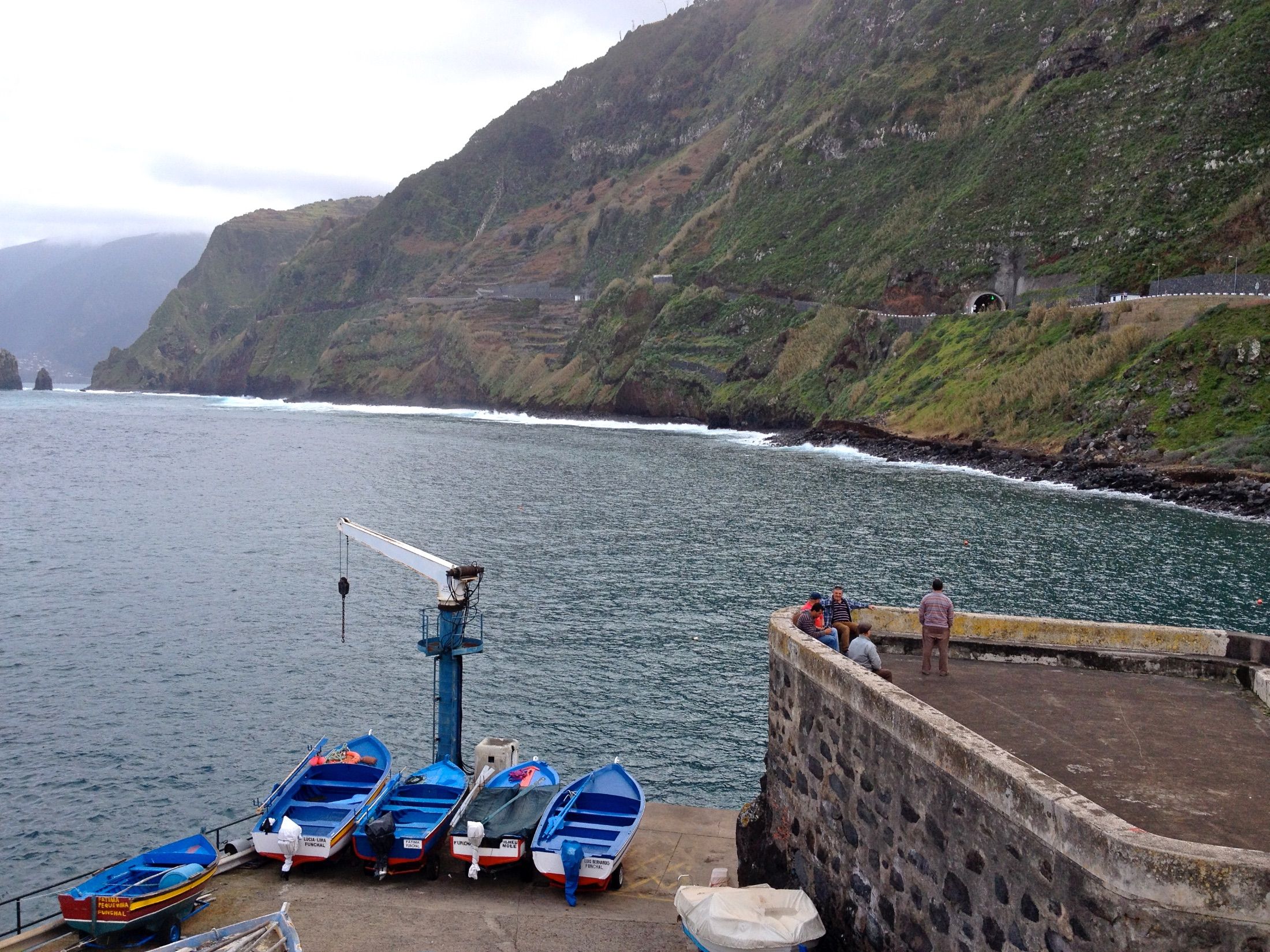 Barcos de pesca azuis junto a uma grua portuária, com arribas íngremes do outro lado da baía