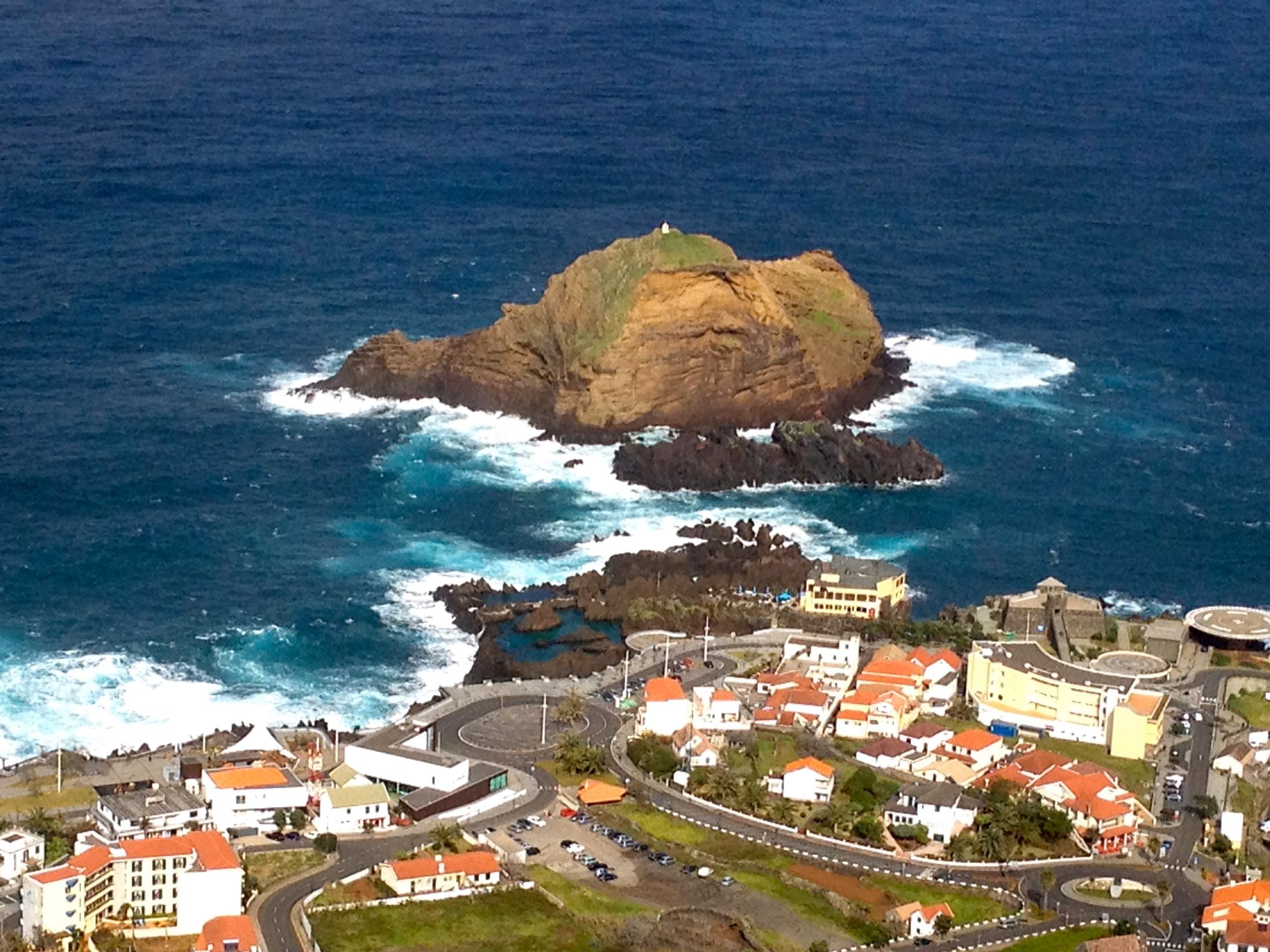 Vila costeira com edifícios de telhados vermelhos, piscinas entre rochas e um ilhéu rodeado de rebentação