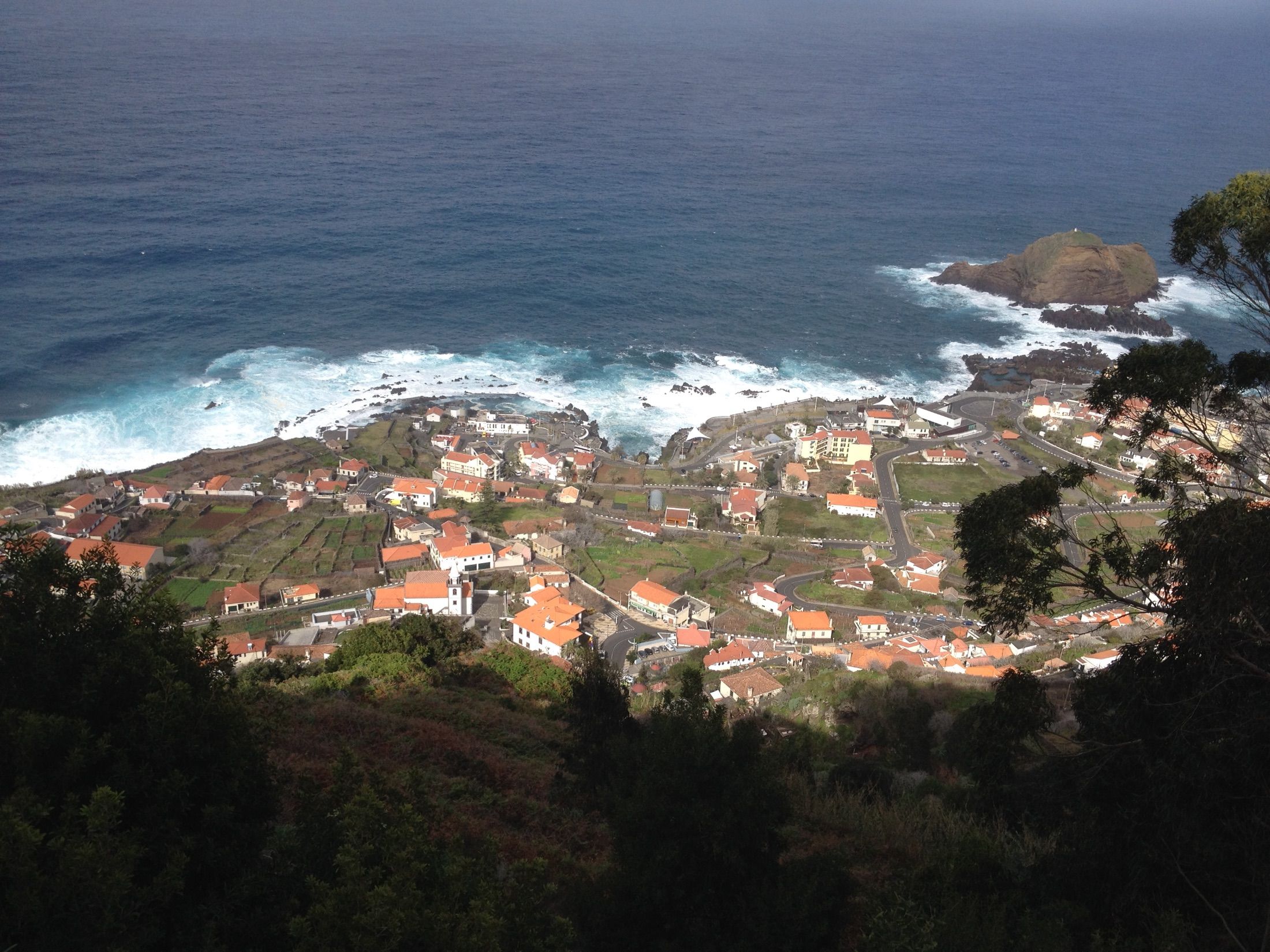 Vista elevada do Porto Moniz, com edifícios de telhados vermelhos, costa rochosa e rebentação