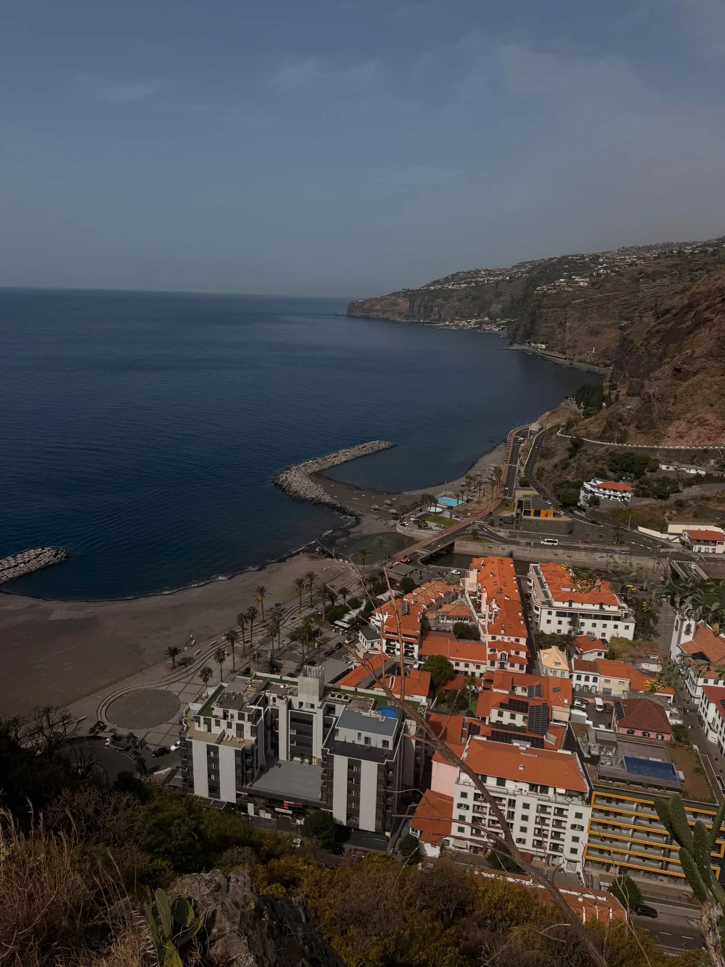 Ribeira Brava from the hillside above: orange-roofed town, palm-lined promenade, dark-sand beach and breakwater, with the south coast cliffs beyond
