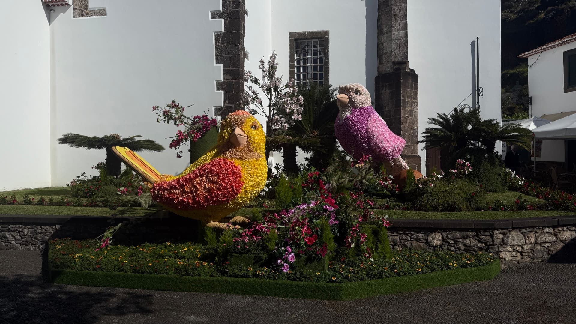 Pássaros gigantes amarelo e cor-de-rosa feitos de flores junto à igreja branca de Santa Cruz, Madeira