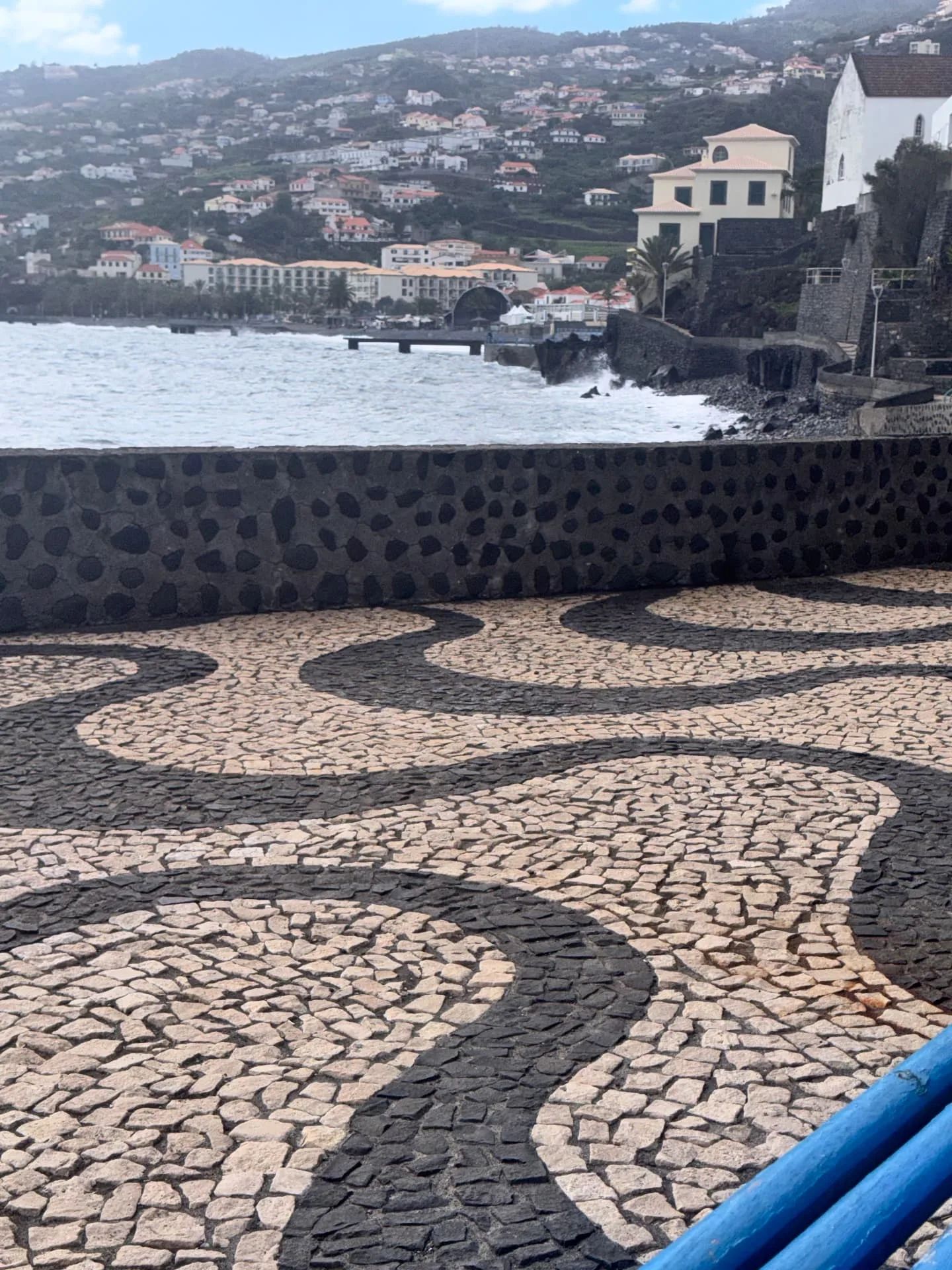 Calçada preta e branca junto à muralha com mar agitado e hotéis de Santa Cruz ao fundo