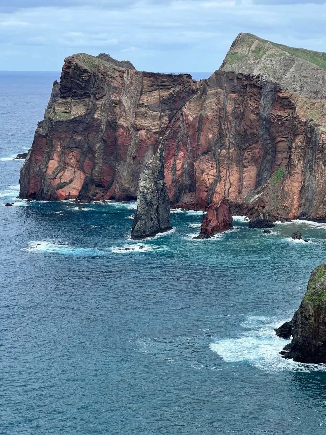 Vista de perto das falésias em camadas vermelhas e cinzentas, com um farol solitário na Ponta de São Lourenço
