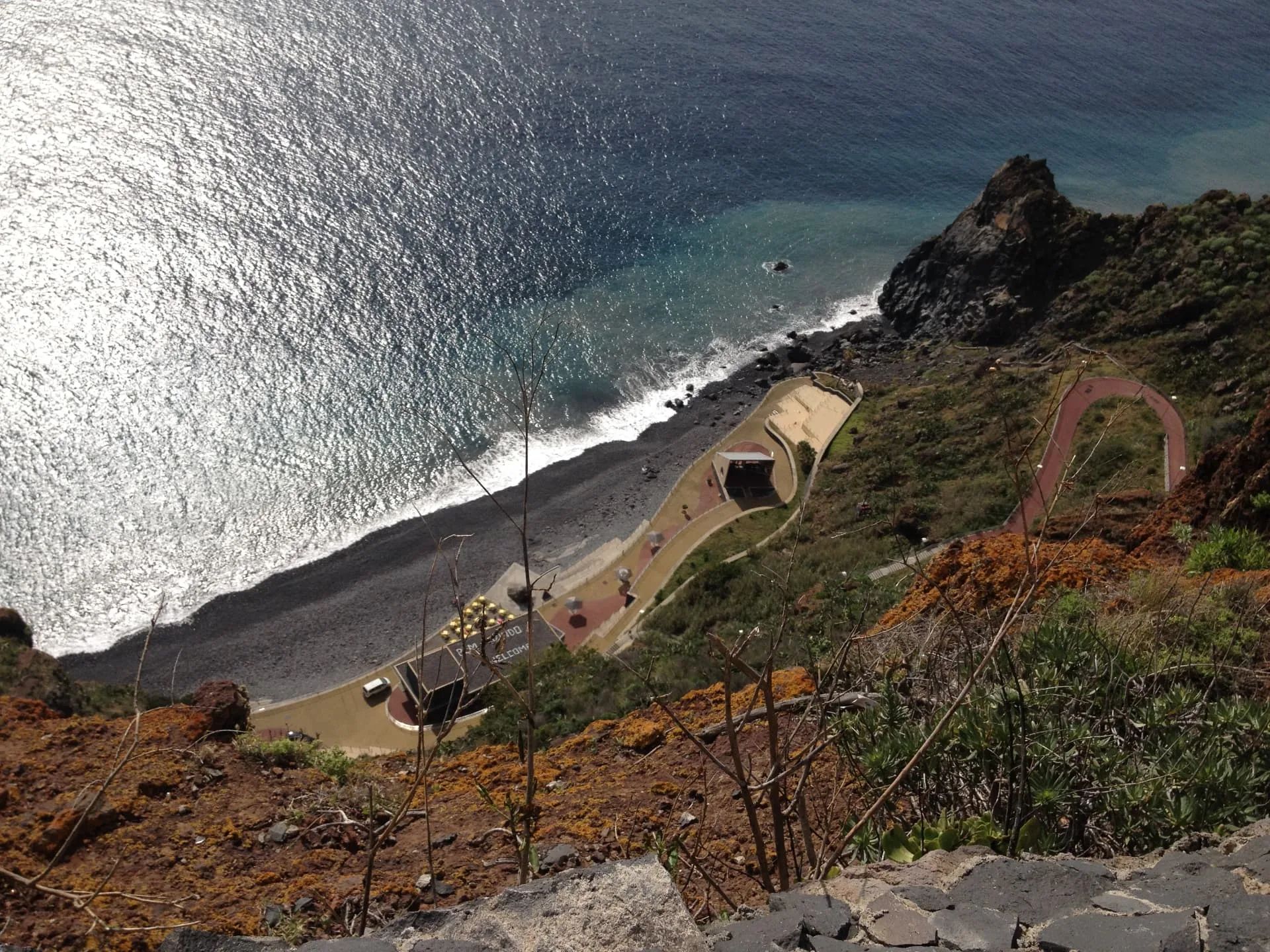 Looking straight down from the Cristo Rei clifftop to Praia do Garajau pebble beach, promenade and yellow umbrellas