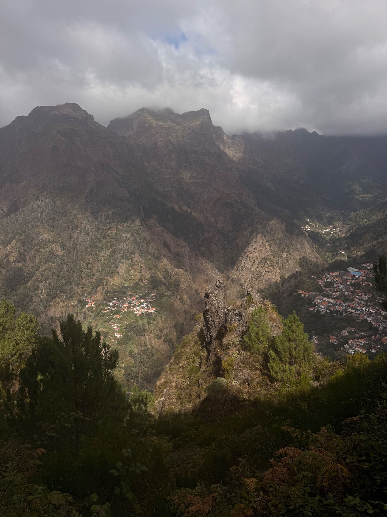 Steep mountain valley with red-roofed houses, foreground pines and low cloud over rocky peaks